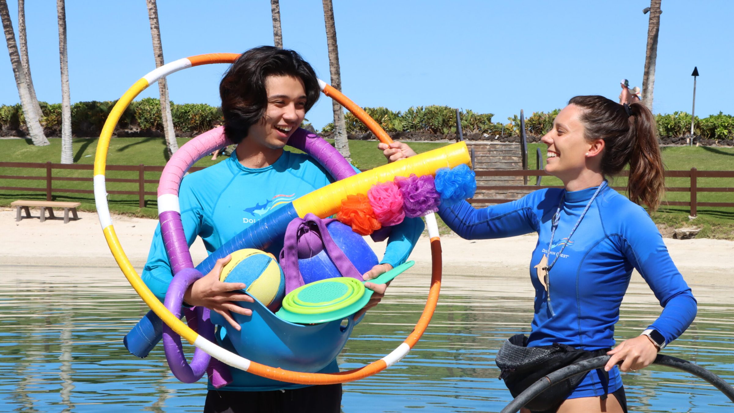 A couple in blue swimsuits holding a hula hoop, with dolphin toys in the pool.