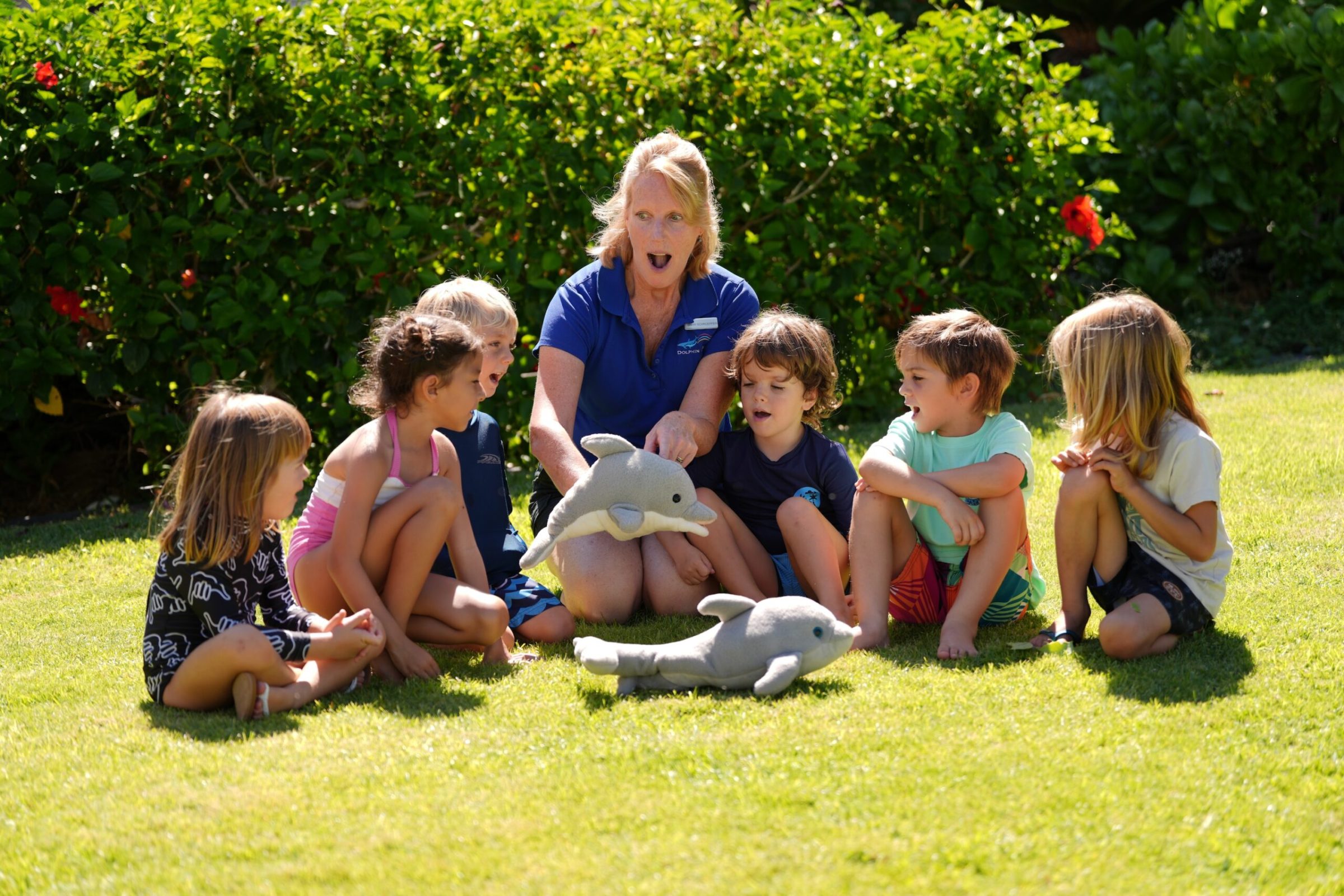A group of young children sitting on the grass together with their teacher holding a dolphin stuff toy. A group of young children sitting on the grass together with their teacher holding a dolphin stuff toy.
