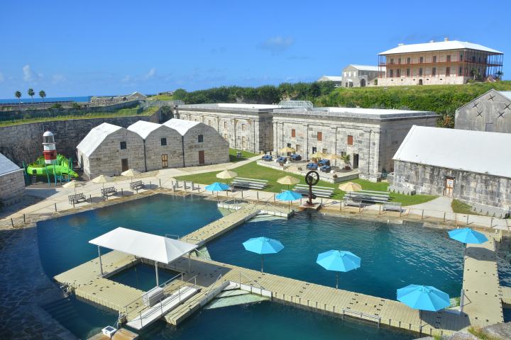 Historic fort with stone buildings, pools with blue umbrellas, and a playground slide, under a clear blue sky.