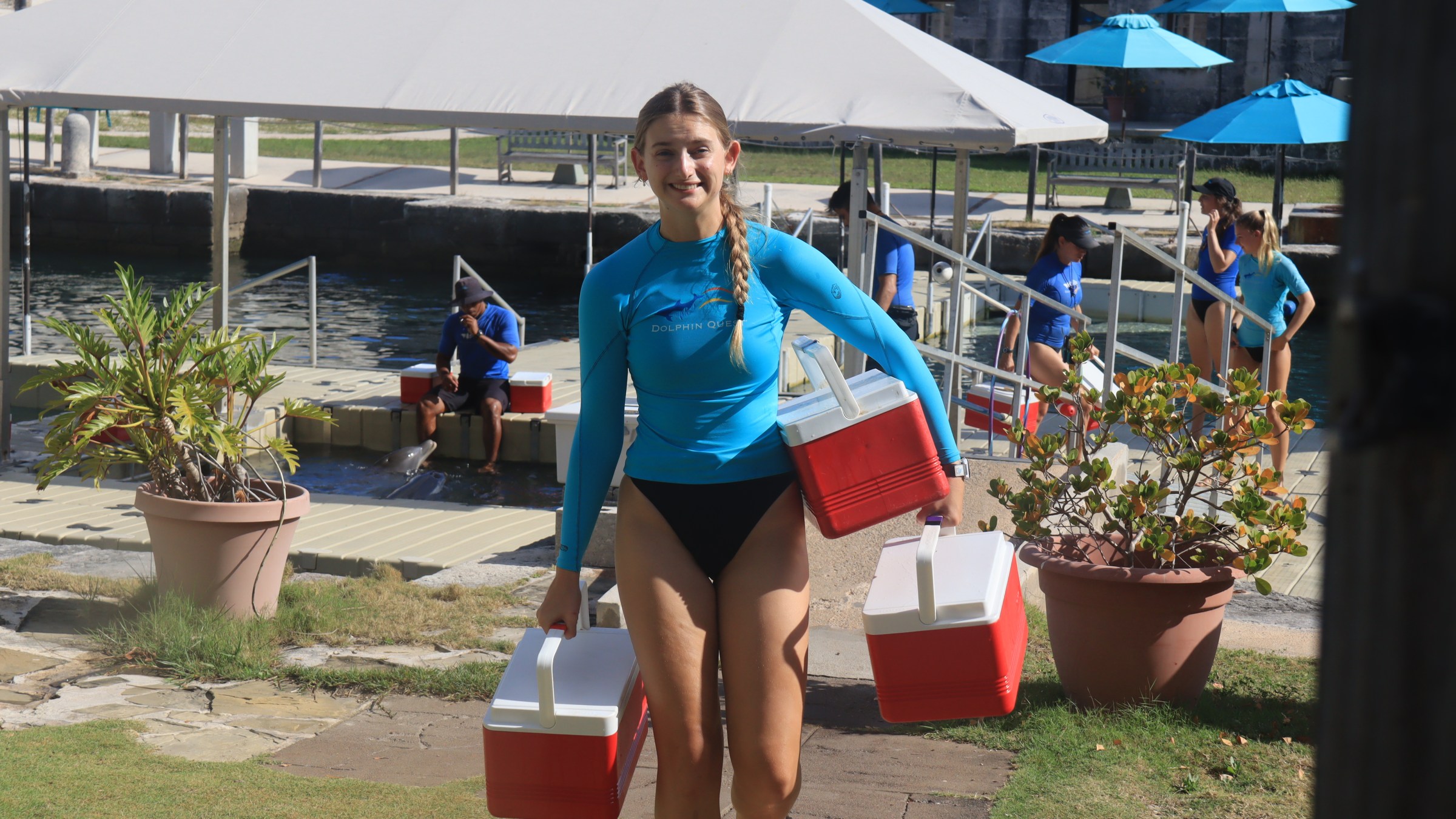 Person in blue wetsuit carrying two red coolers near a pool with other people and umbrellas.