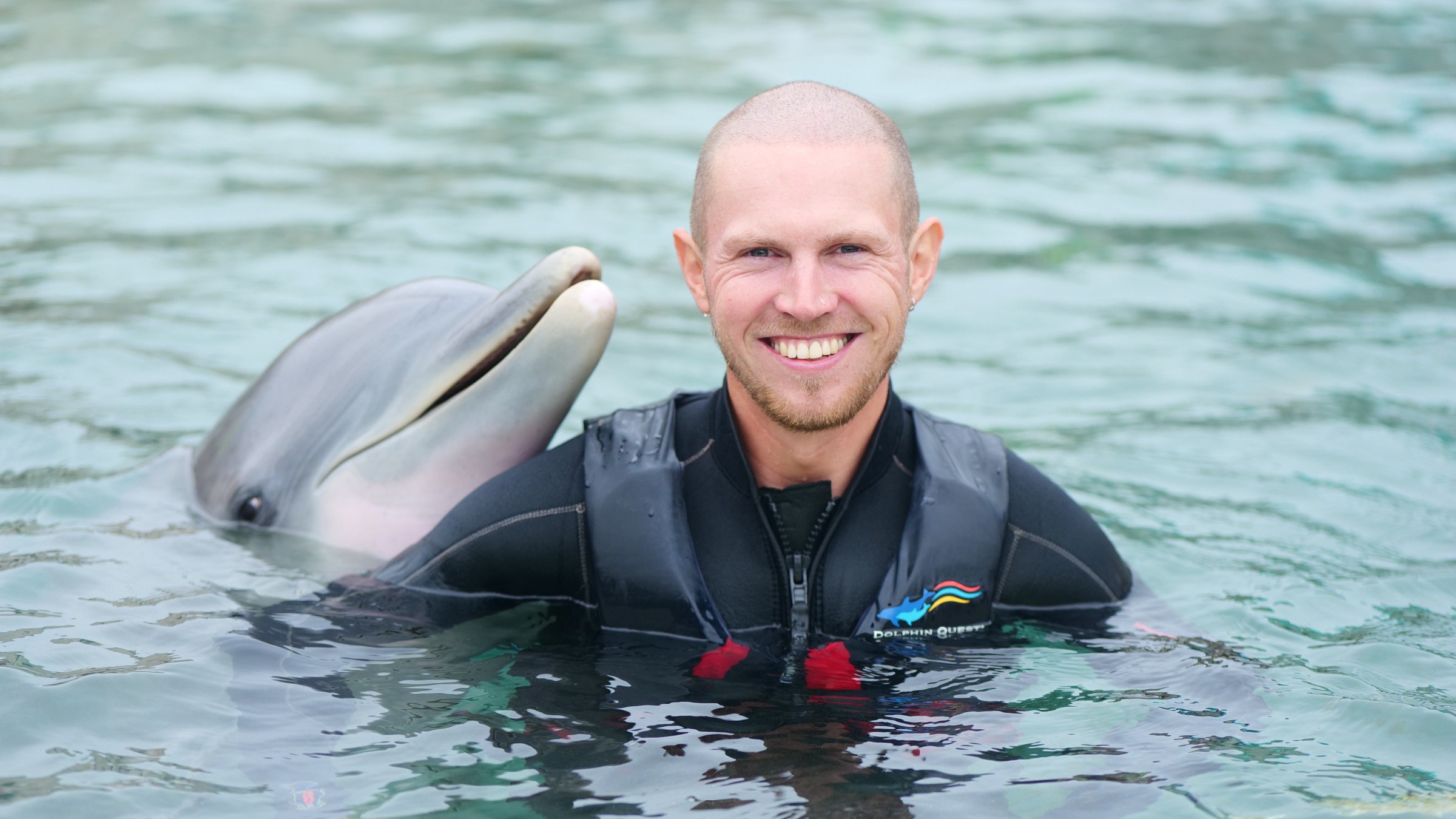Person in a wetsuit smiling with a dolphin in water.