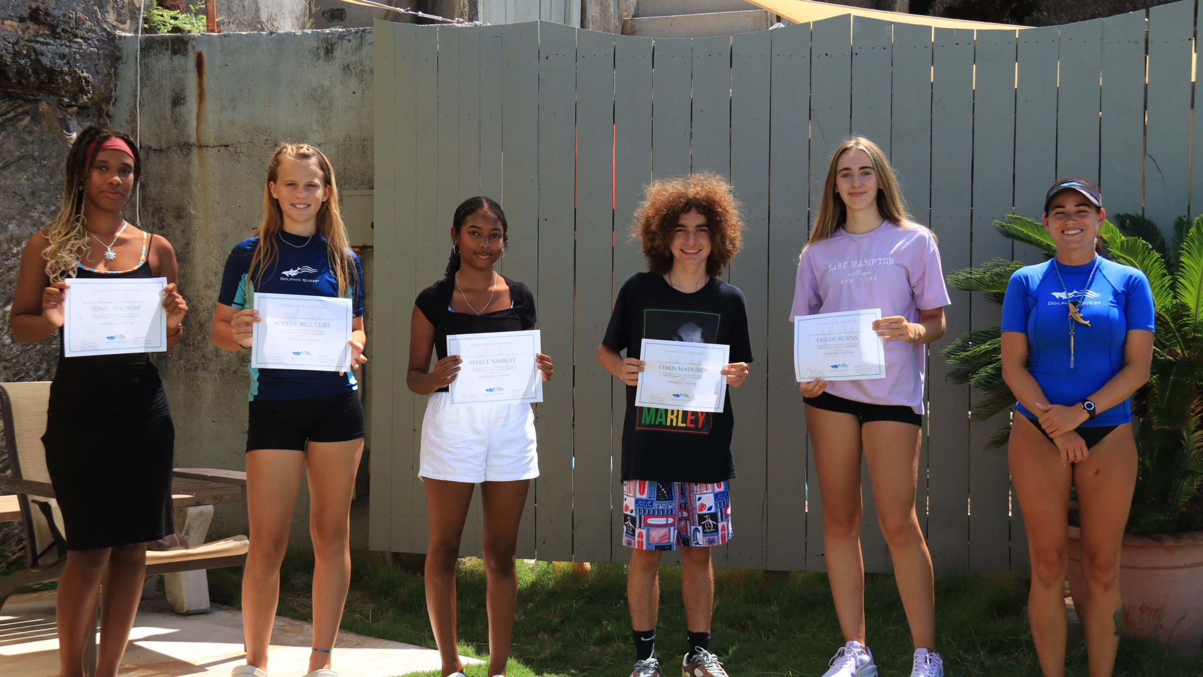 Five people standing outside holding certificates, with a sixth person in blue attire smiling beside them.