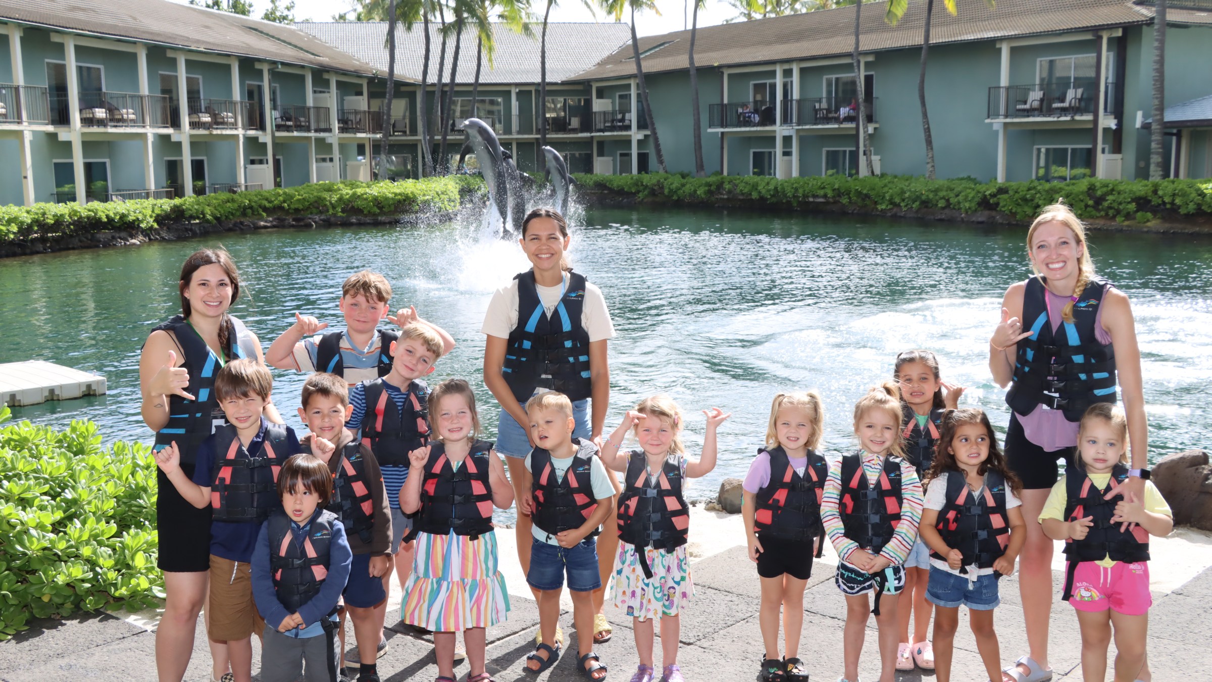 Group of children and adults in life vests posing by water with dolphins in the background.